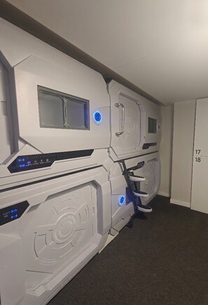 A room with a large, white, futuristic-looking machine with a control panel and a small window. There is a door with the numbers '17' and '18' on it.