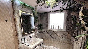 Baño con lavamanos de piedra, espejo y ventana con artículos de aseo.
