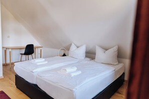 Ein Doppelbett mit weißer Bettwäsche und Kissen, ein Schreibtisch mit Stuhl sowie ein Nachttisch mit Lampe.
