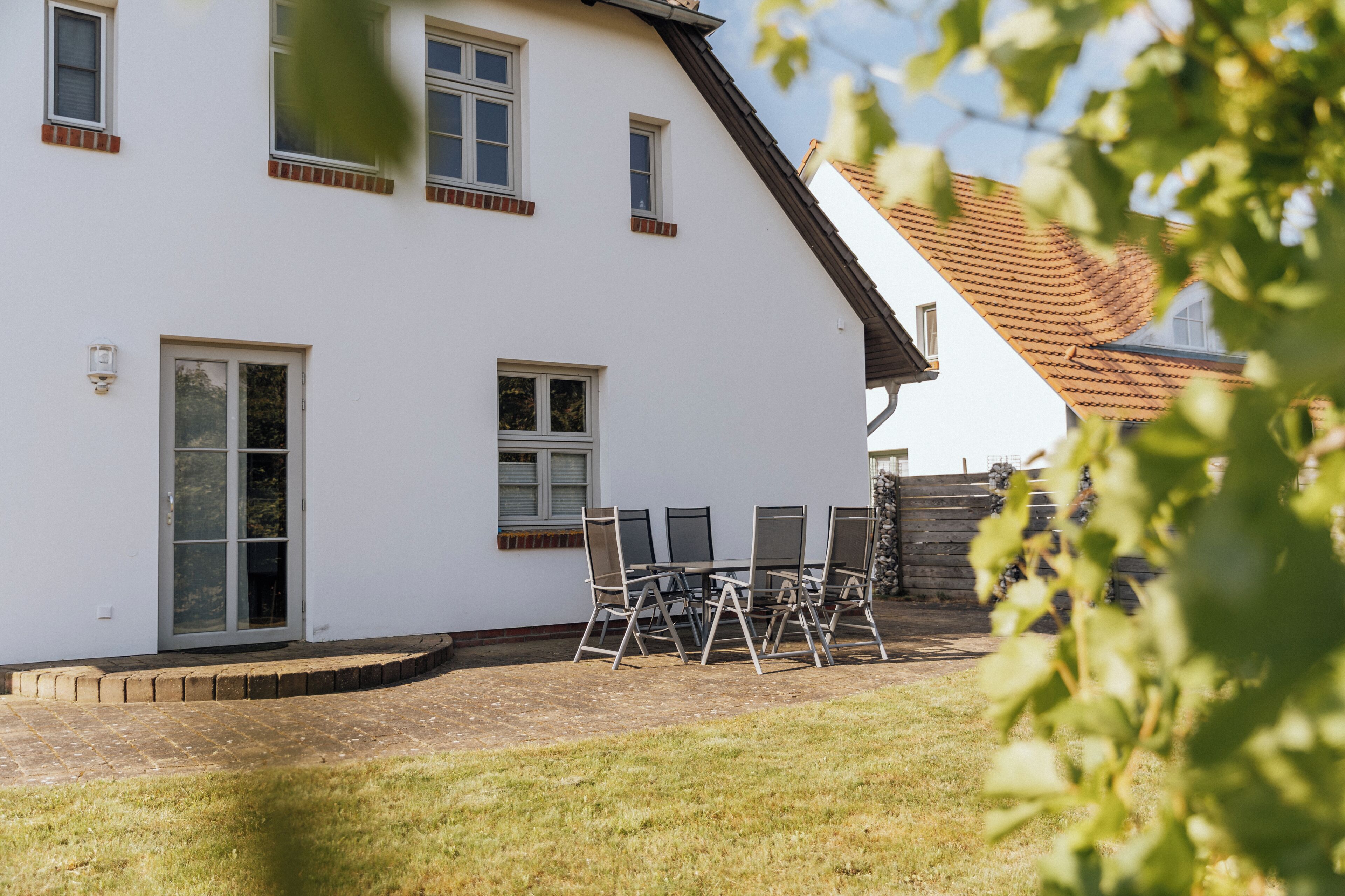 A white building with a sloped roof, a door, and windows. There is a patio with chairs and a table.