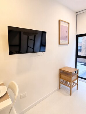 Ein modernes Zimmer mit an der Wand befestigtem Fernseher, einem kleinen Holzregal, einem weißen Stuhl und einem gerahmten Bild.