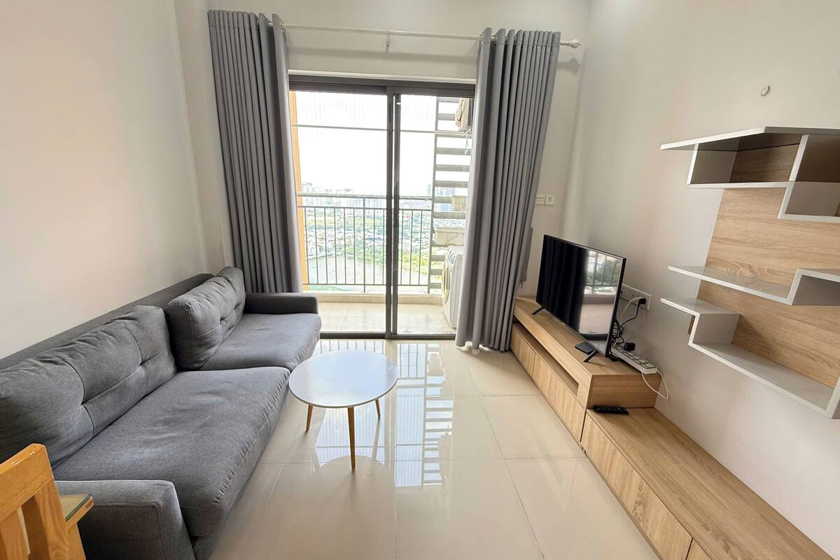 Ein modernes Wohnzimmer mit grauem Sofa, einem kleinen runden Tisch, einem Fernseher auf einem Holzschrank und einem Balkon mit grauen Vorhängen.