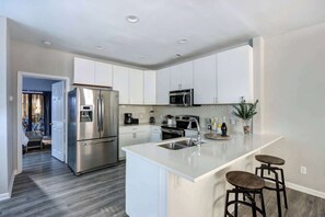 A modern kitchen with stainless steel appliances, a central island with bar stools, and a view into the adjacent living area. - The Garden @ Hunter’s Knoll | Walk to Forsyth Park (Savannah)