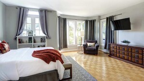 Ein Schlafzimmer mit einem Bett, einem Sessel, einem Fernseher, einem Schminktisch und einem Fenster mit Vorhängen.