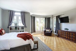 Ein Schlafzimmer mit einem Bett, einem Sessel, einem Fernseher, einem Schminktisch und einem Fenster mit Vorhängen.