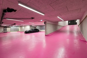 Un parking au sol recouvert d’un revêtement et aux murs de couleur rose, avec une voiture noire et des piliers en béton.