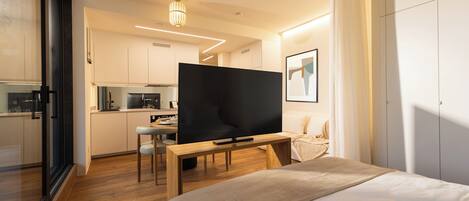 A modern hotel room with a large bed, a television, a dining area, and a kitchenette.