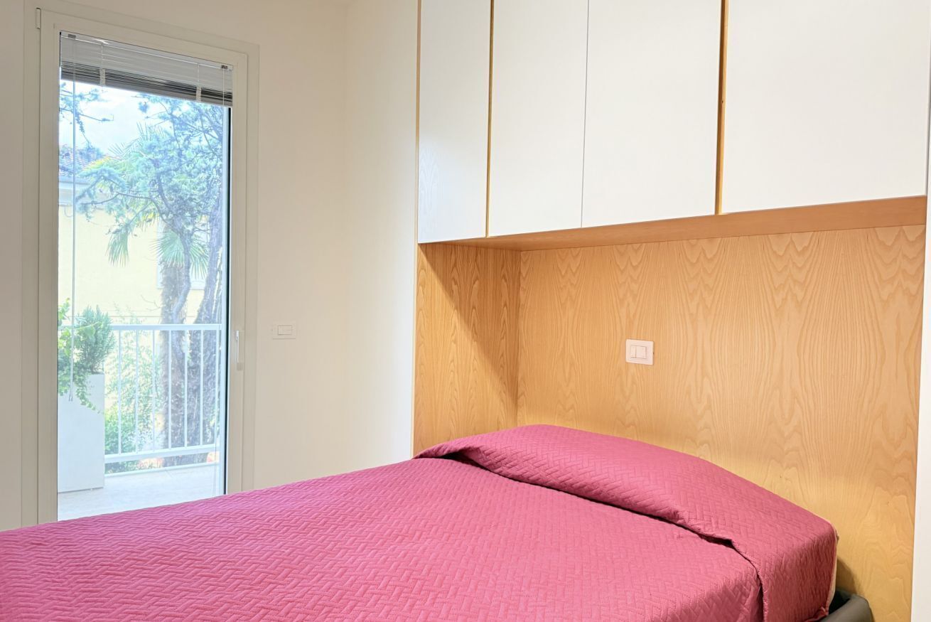 A bedroom with a bed, a wooden headboard, a window with blinds, and a view of greenery outside.