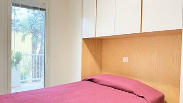 A bedroom with a bed, a wooden headboard, a window with blinds, and a view of greenery outside.