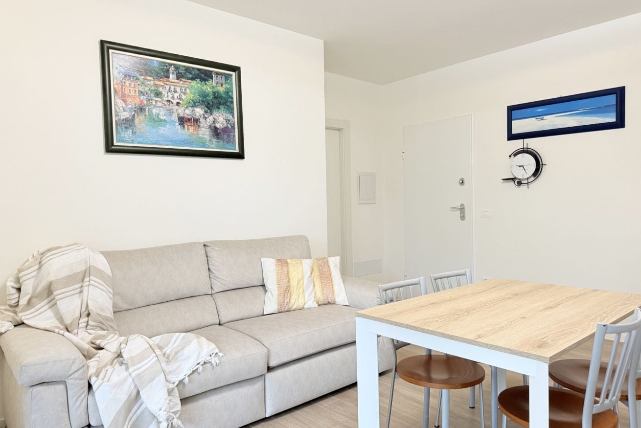 Ein Wohnzimmer mit einer Couch, einem Holzesstisch und Stühlen. An der Wand hängt ein gerahmtes Bild und eine Uhr.