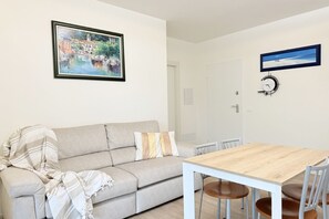 Ein Wohnzimmer mit einer Couch, einem Holzesstisch und Stühlen. An der Wand hängt ein gerahmtes Bild und eine Uhr.
