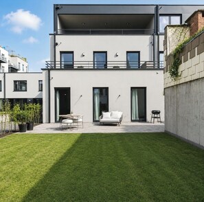 Ein modernes, zweistöckiges Gebäude mit flachem Dach, großen Fenstern und einer überdachten Terrasse mit weißem Sofa und Tisch.