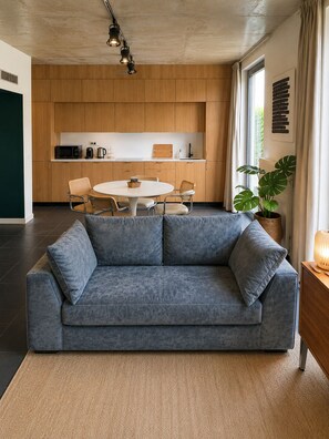Ein modernes Wohnzimmer mit grauem Sofa, einem runden Esstisch und einer Küche im Hintergrund.