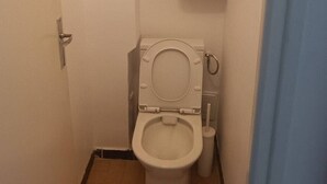 Een kleine badkamer met toilet, ventilatie en opslagplanken voor toiletartikelen.