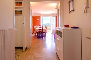 Un couloir mène à une salle à manger avec une table et des chaises, une cuisine équipée d’un réfrigérateur, et une porte donnant sur l’extérieur.