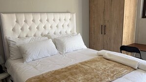 A bedroom with a tufted headboard, white bedding, a large wooden wardrobe, and a chair.