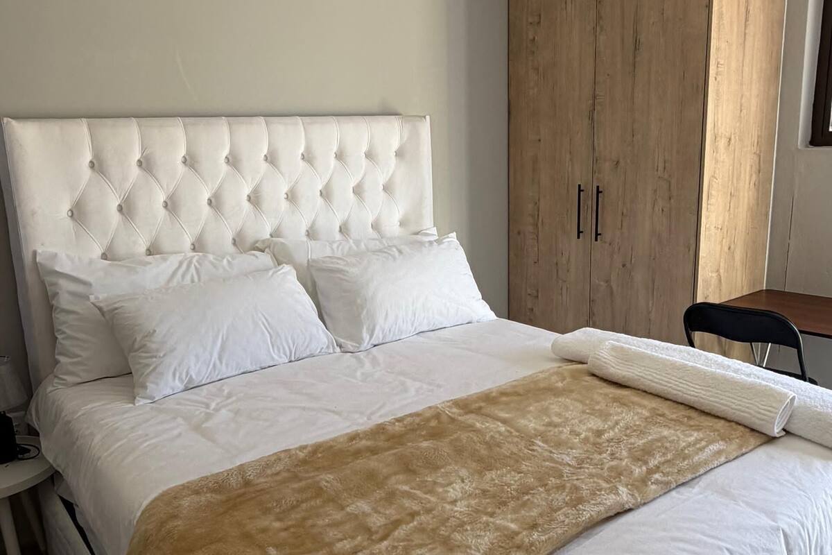 A bedroom with a tufted headboard, white bedding, a large wooden wardrobe, and a chair.