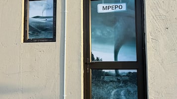 Un edificio con un letrero que dice 'MPEPO' sobre una puerta, una pequeña ventana con una imagen de un paisaje, y un accesorio de iluminación en la pared.
