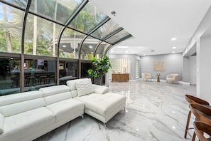 Ein modernes Wohnzimmer mit einem Ecksofa, einem Essbereich mit einem Glastisch und Blick auf Palmen durch große Fenster.