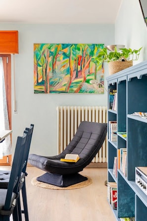 Une pièce comprenant un fauteuil de couleur noire, une bibliothèque, un radiateur et un tableau coloré accroché au mur.
