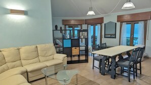 Una sala de estar con sofá, una mesa de centro de vidrio, una mesa de comedor con sillas, una estantería y vistas a través de las ventanas.