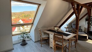 A room with a sloped ceiling, a wooden desk, a chair, a potted plant, and a view of a town through a large window.