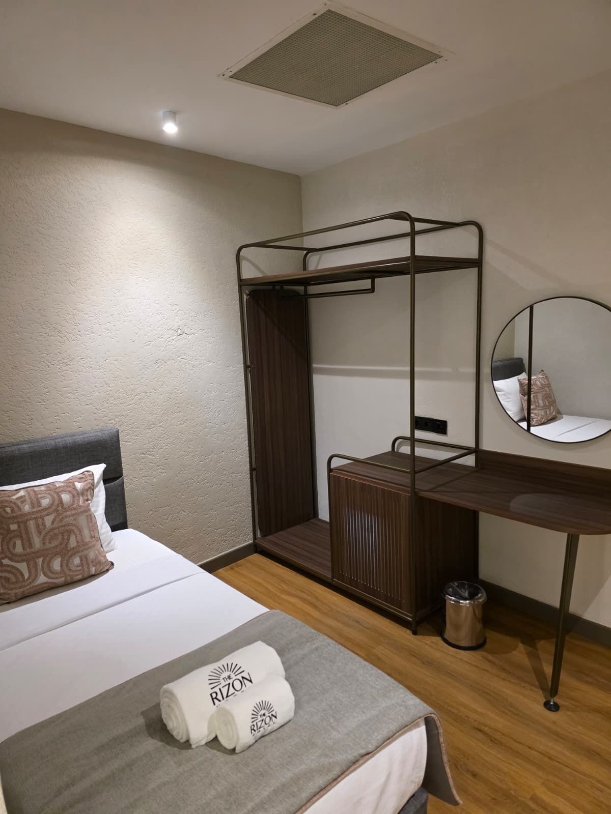 A compact hotel room with a bunk bed, a desk, a small trash bin, and a round mirror on the wall.