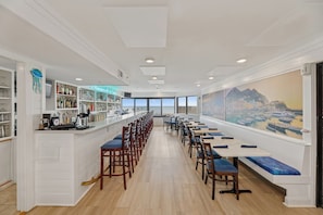 Une longue salle à manger avec de grandes tables et des chaises, un bar à gauche et une grande fenêtre donnant sur le paysage urbain.