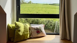 Ein Zimmer mit Fensterblick auf ein grünes Feld, eine Holzbank mit Kissen und eine weiße Wand.
