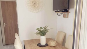 Ein Essbereich mit Holztisch, vier Stühlen, einer Topfpflanze, einem gewebten Korb und einem an der Wand befestigten Fernseher.