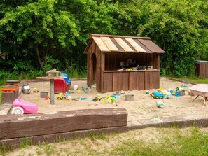 Ein Spielhäuschen aus Holz mit geneigtem Dach, umgeben von einer Sandspielplatzfläche mit verschiedenen Spielsachen und Geräten.