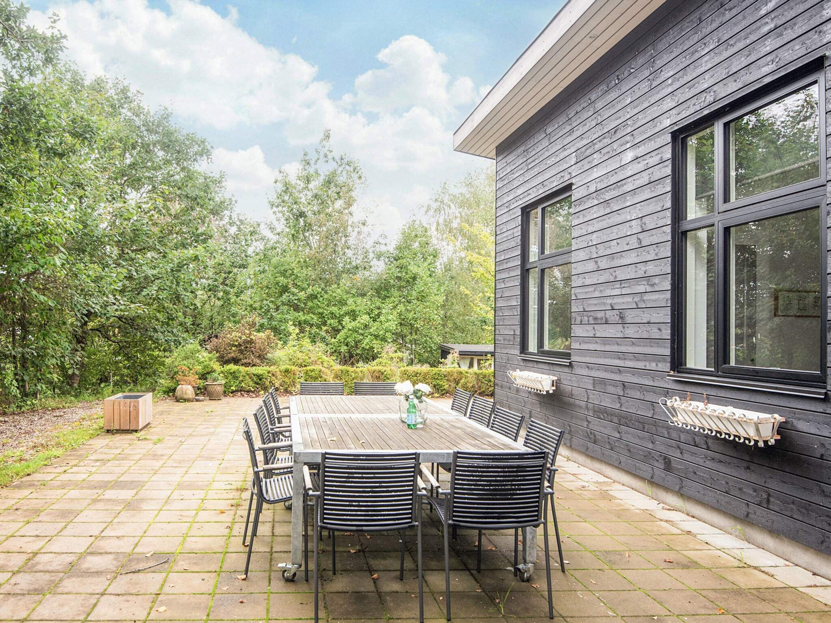 A patio with a wooden table and chairs, surrounded by a garden and a building with large windows.