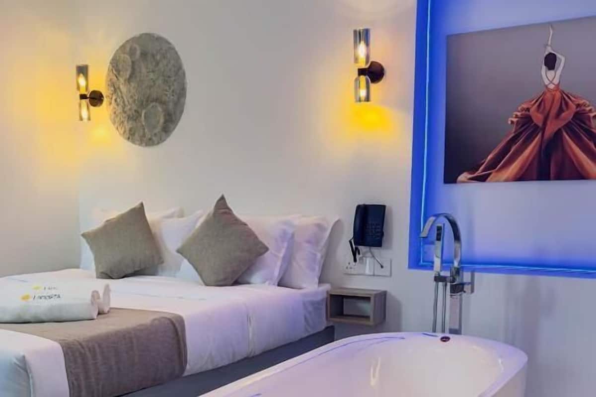 Quarto de hotel moderno com uma banheira grande e independente, cama com roupa de cama branca, mesa de cabeceira e iluminação embutida na parede.