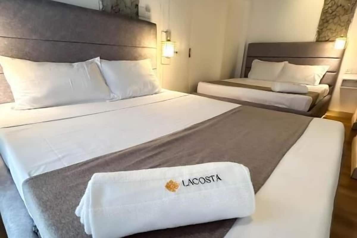 Quarto de hotel com duas camas, uma parede de destaque em pedra e uma toalha com a marca 'LACOSTA'.