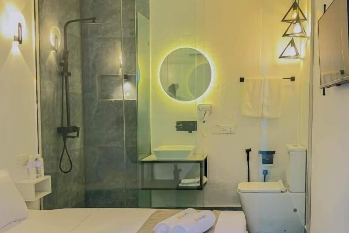 Quarto de hotel moderno com cama, chuveiro em box de vidro, espelho redondo, pia e vaso sanitário.