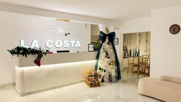 A hotel reception area with a modern design, a reception desk, a large mirror, and a seating area with a sofa. - La Costa Hotel (Seeduwa)