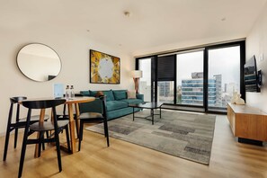 Ein modernes Wohnzimmer mit einem runden Spiegel, einem grünen Sofa, einem Couchtisch und Blick auf die Stadt durch große Fenster.