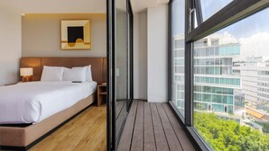Ein modernes Hotelzimmer mit einem großen Bett, einem Nachttisch, einer Lampe und Blick auf die Stadt durch große Fenster.