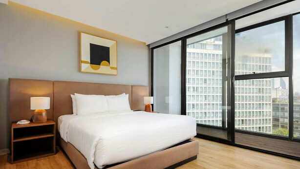 Ein modernes Hotelzimmer mit einem großen Bett, zwei Nachttischen mit Lampen, einem gerahmten Kunstwerk über dem Bett und einem großen Fenster mit Blick auf die Stadt.