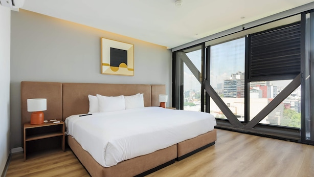 Una habitación de hotel moderna con una cama grande, vistas a la ciudad a través de ventanales del piso al techo y una obra de arte enmarcada en la pared.