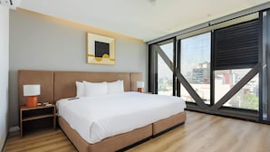 Una habitación de hotel moderna con una cama grande, una mesita de noche, una lámpara, un cuadro enmarcado y un amplio ventanal con vistas a la ciudad.