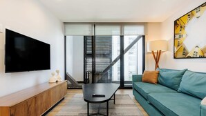 Una sala de estar moderna con un televisor de pantalla plana, un mueble de madera, una mesa negra, un sofá color turquesa y un ventanal con vistas a edificios.