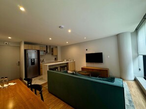 Eine moderne Küche mit integriertem Kühlschrank, Mikrowelle und Herd. Es gibt eine Essecke mit Holztisch und Stühlen, sowie ein grünes Sofa mit Kissen. Ein Flachbildfernseher ist an der Wand befestigt.