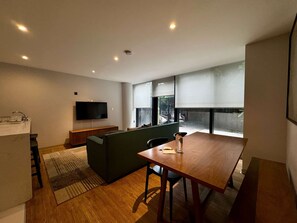 Ein modernes Wohnzimmer mit einem Holzesstisch, einem grünen Sofa, einem Flachbildfernseher und einer Küchenzeile mit Spüle.