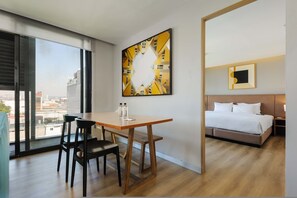 Ein Hotelzimmer mit großem Fenster, einem Bett, einem Tisch mit Stühlen und einem Gemälde an der Wand.