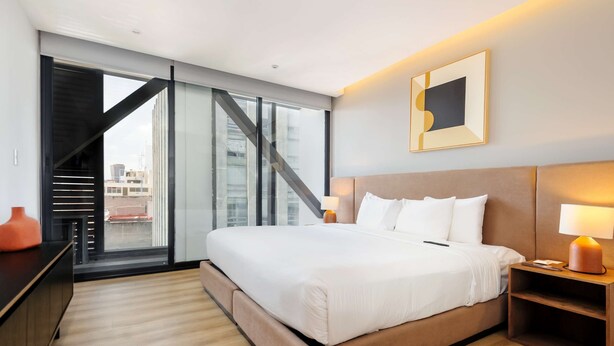 A modern hotel room with a large bed, a desk, a chair, a lamp, and a view of the city through large windows. - w Amazing 1BR w/ AC in the Heart of Roma Sur (Mexico City)