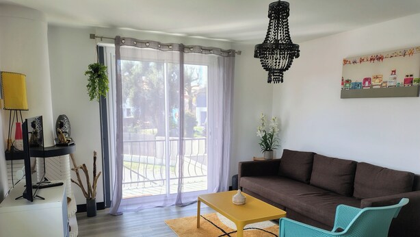 A living room with a brown sofa, a yellow table, a turquoise chair, a chandelier, and a view through a glass door. - Spacious apartment near ocean and shops (Angresse)