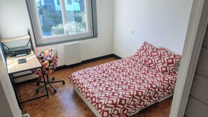 A bedroom with a bed, a desk with a chair, a window, and a radiator. - Spacious apartment near ocean and shops (Angresse)
