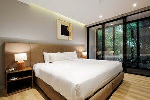 Ein modernes Hotelzimmer mit einem großen Bett, Nachttischen, einem kleinen Tischchen, einem gerahmten Bild an der Wand und einem großen Fenster mit Blick auf Bäume.