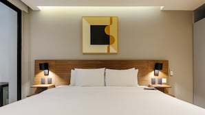 Quarto de hotel com uma cama grande, cabeceira de madeira, duas mesas de cabeceira com luminárias, uma obra de arte pendurada na parede e uma janela.